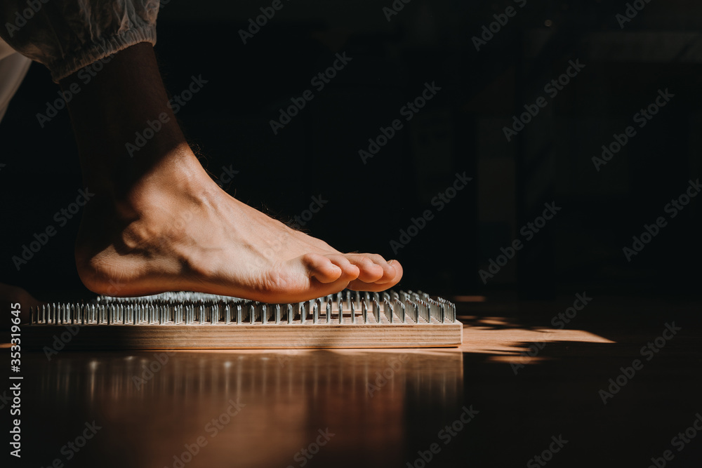 Close-up of unrecognizable brave man in white pants standing barefeet on bed of nails at morning yoga practice. Balance exercise, courage and patience. Close-up of unrecognizable brave man in white pants standing barefeet on bed of nails at morning yoga practice. Balance exercise, courage and patience.