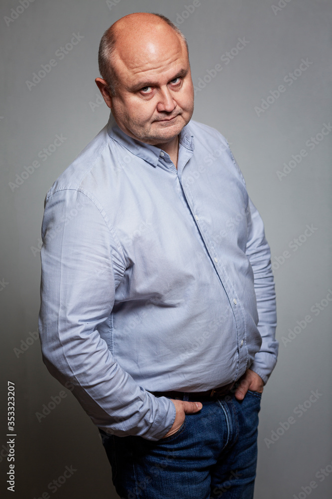 Adult bald serious man in this shirt and jeans on a gray background. Looks strictly at the camera. Vertical. Adult bald serious man in this shirt and jeans on a gray background. Looks strictly at the camera. Vertical.