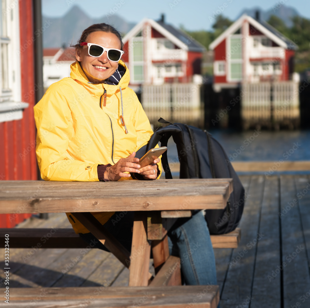Young woman in a yellow jacket on a background of traditional red Norwegian rorbu houses with a smartphone in hand, traveling to Norway Lofoten Islands Young woman in a yellow jacket on a background of traditional red Norwegian rorbu houses with a smartphone in hand, traveling to Norway Lofoten Islands
