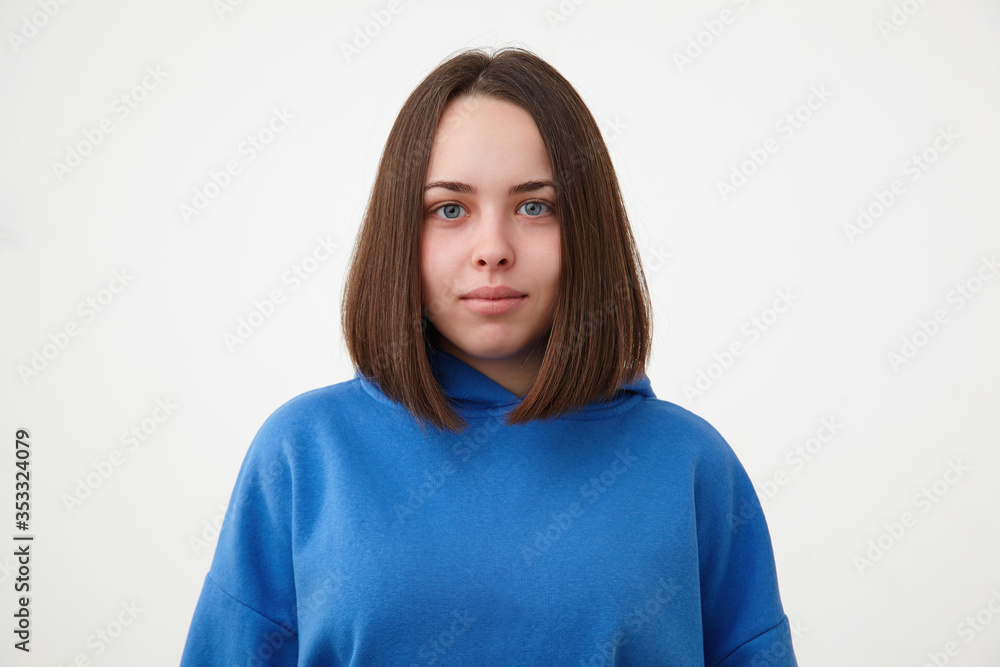 Portrait of young beautiful blue-eyed brunette lady with short haircut looking positively at camera with light smile, standing over white background in sporty wear