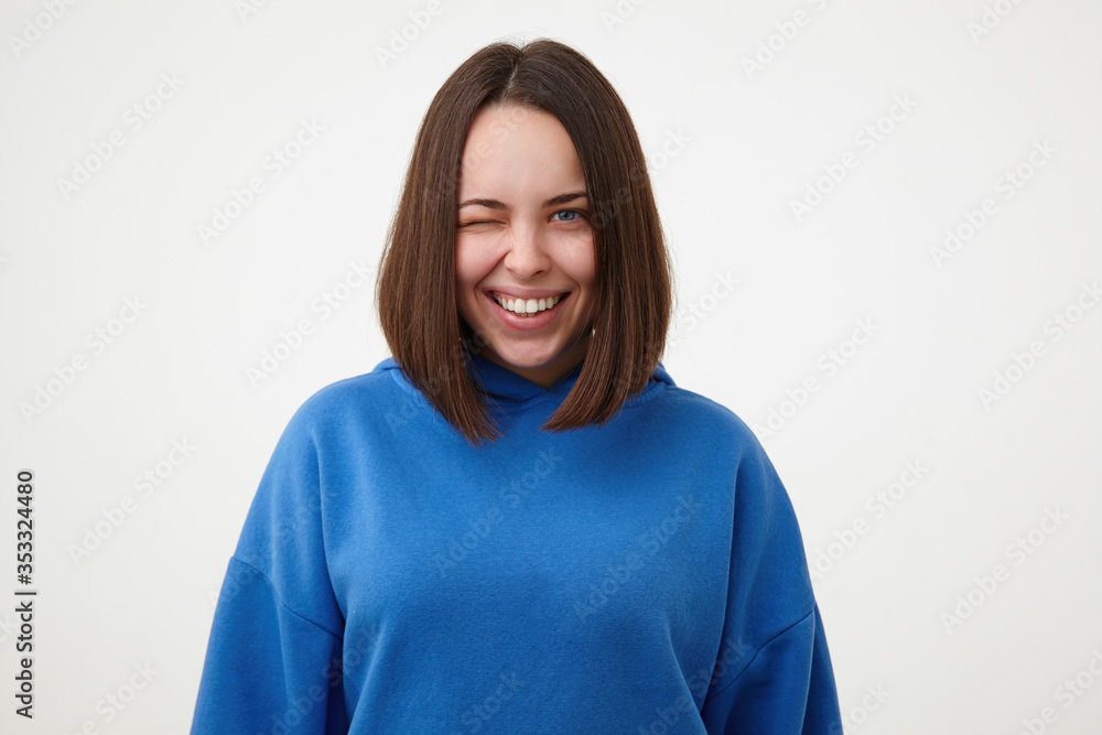 Cheerful young attractive short haired blue-eyed woman with bob haircut smiling gladly while winking at camera, dressed in blue hoodie while posing over white background Cheerful young attractive short haired blue-eyed woman with bob haircut smiling gladly while winking at camera, dressed in blue hoodie while posing over white background