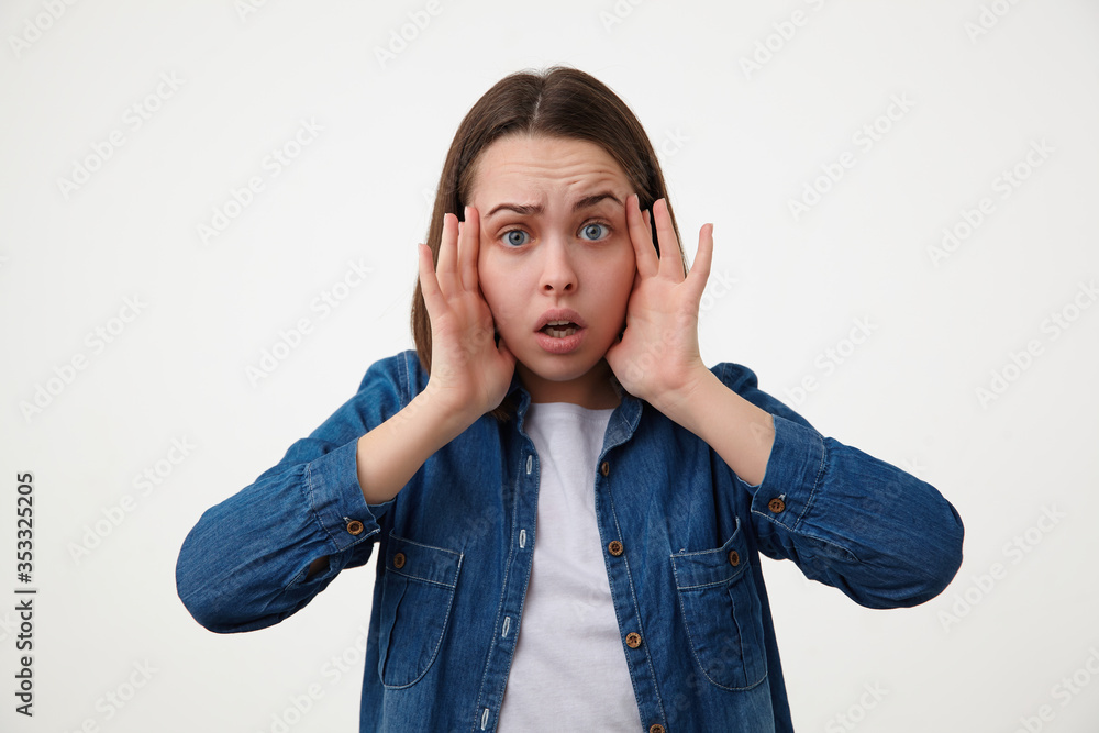 Shocked young brown haired blue-eyed lady without makeup holding her face with raised hands while looking scaredly at camera, isolated over white background Shocked young brown haired blue-eyed lady without makeup holding her face with raised hands while looking scaredly at camera, isolated over white background