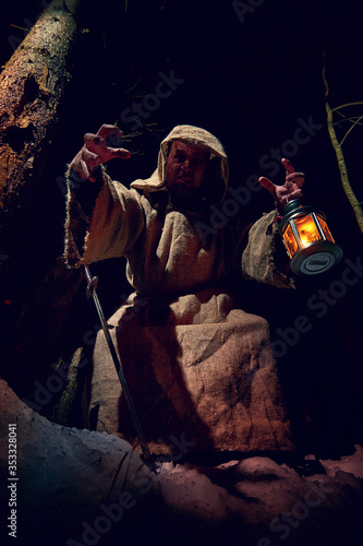 Terrible medieval monk in canvas sackcloth robe with lattern in dark forest and red light of moon on winter night. Fantasy or fairy tale about wandering monk. Concept of forces of good and evil