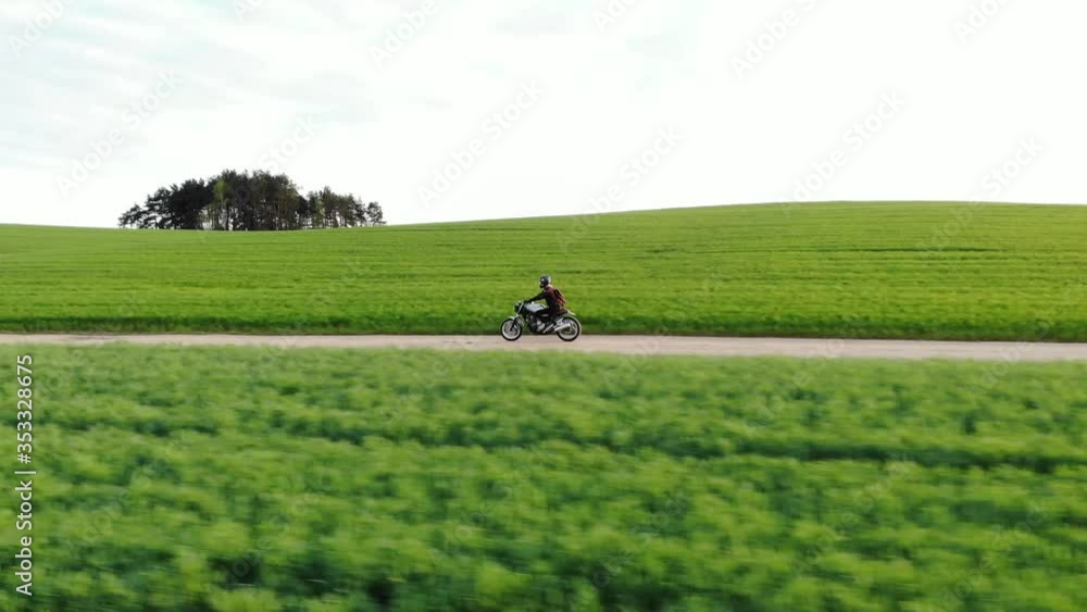 Classic custom motorcycle rides on a village road through a green field. Drone video in 4k