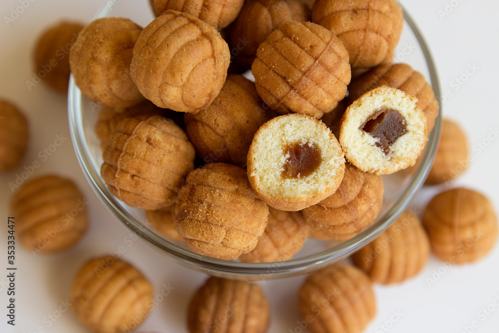 Round ruddy biscuit delicacy dessert in the form of small balls with boiled caramel inside. Cookies with filling are located in a transparent glass bowl and are scattered nearby on a white table. Round ruddy biscuit delicacy dessert in the form of small balls with boiled caramel inside. Cookies with filling are located in a transparent glass bowl and are scattered nearby on a white table.