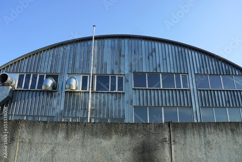 facade of an industrial building from a gray metal wall with windows and fan pipes behind a concrete fence against a blue sky © butus
