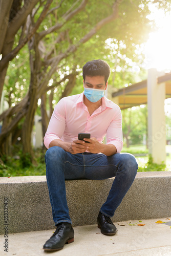 Full body shot of young Indian businessman with mask using phone and sitting at the park