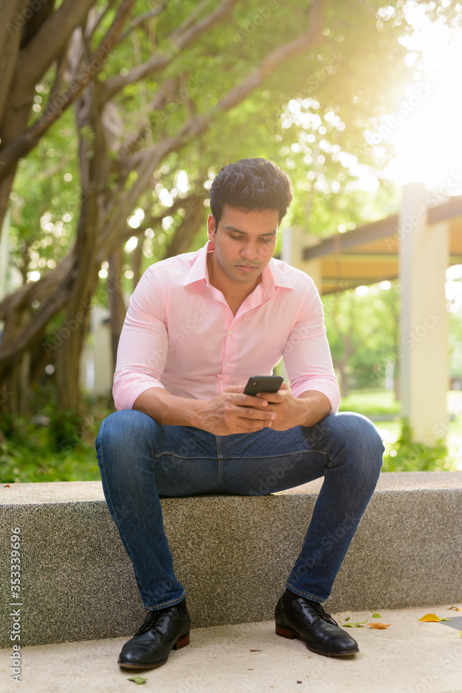 Full body shot of young handsome Indian businessman using phone and sitting at the park Full body shot of young handsome Indian businessman using phone and sitting at the park