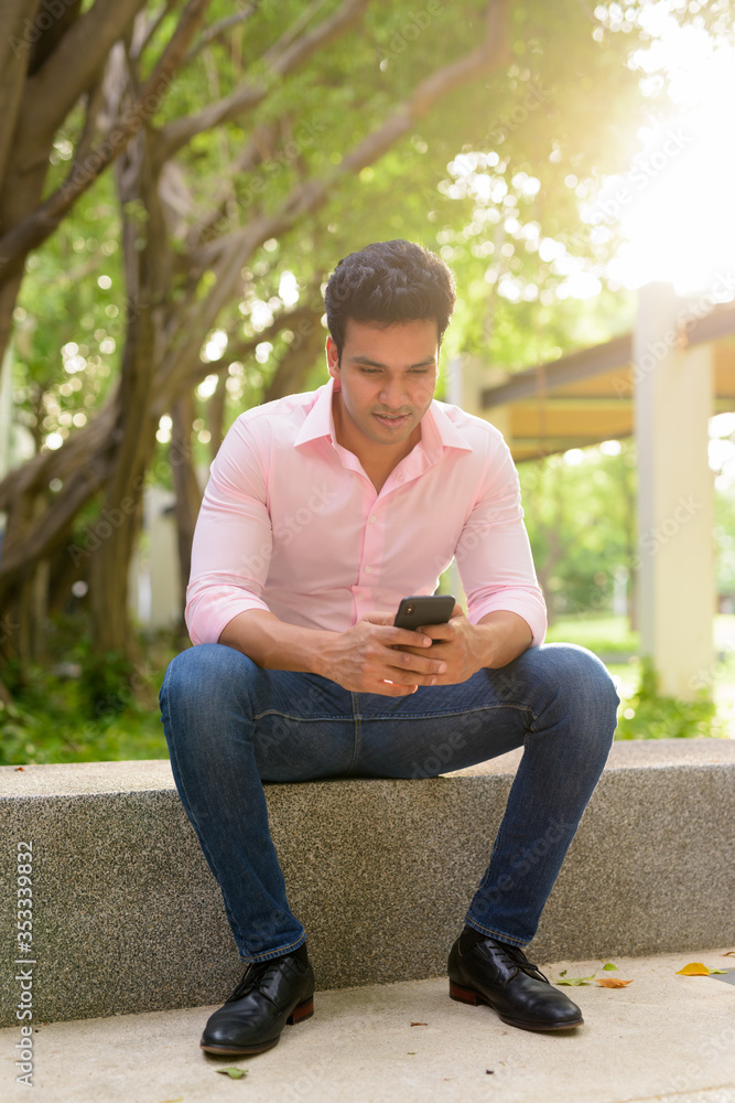 Full body shot of happy young handsome Indian businessman using phone and sitting at the park Full body shot of happy young handsome Indian businessman using phone and sitting at the park