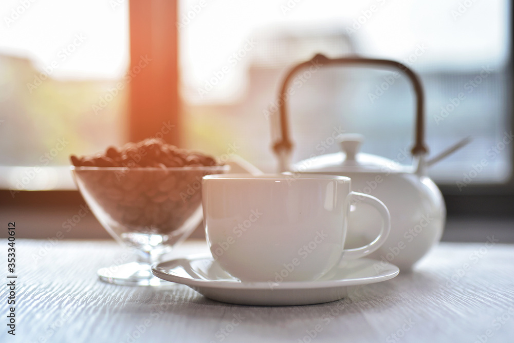 White coffee cup with roasted coffee beans and morning sunrise at the windows background White coffee cup with roasted coffee beans and morning sunrise at the windows background