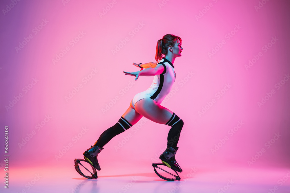 Beautiful redhead woman in sportswear jumping in a kangoo jumps shoes isolated on purple-pink gradient studio background in neon light. Active movement, action, fitness and wellness. Fit female model. Beautiful redhead woman in sportswear jumping in a kangoo jumps shoes isolated on purple-pink gradient studio background in neon light. Active movement, action, fitness and wellness. Fit female model.