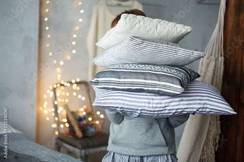 Woman holds stack of grey linen pillows in cozy interiror room with bokeh lights. A stack of pillows hides the face of human.