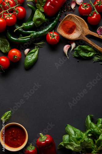 top view of red cherry tomatoes, tomato sauce in bowl, peppercorns, herbs and green chili peppers on black