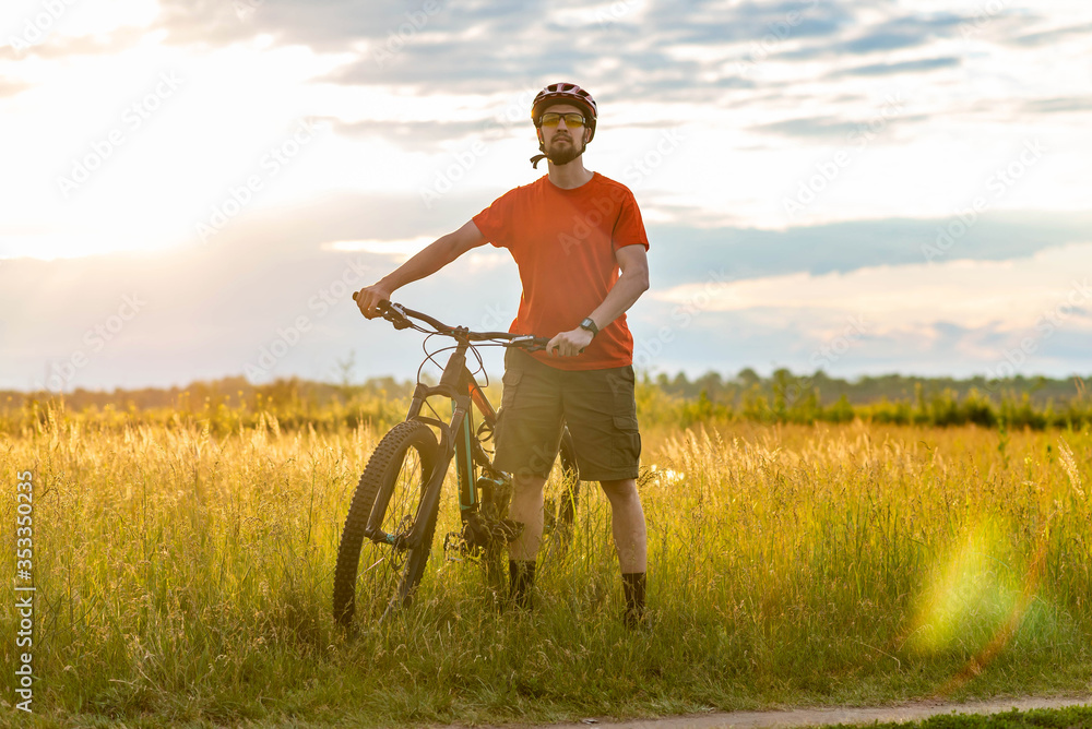 A cyclist in a bright orange T-shirt stands near a bicycle in a meadow during sunset. A cyclist in a bright orange T-shirt stands near a bicycle in a meadow during sunset.