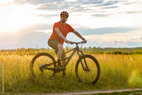 A cyclist in a bright orange T-shirt stands near a bicycle in a meadow during sunset.