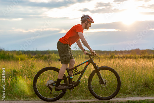 A cyclist in a bright orange T-shirt rides a bicycle along the meadow during sunset.