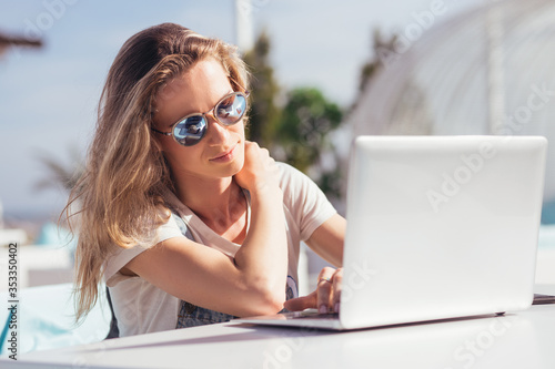 Cute beautiful young woman in sunglasses makes a video call using wireless high-speed Internet while sitting on the veranda by the sea while on vacation on a sunny warm summer day