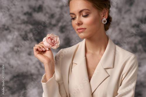 Closeup beauty face portrait of young caucasian woman with perfect makeup and hairstyle. Female model wearing white blazer and holding one pink rose flower photo