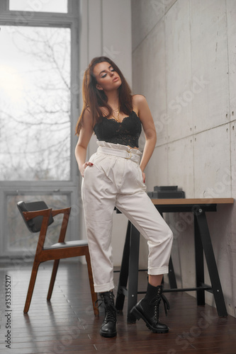 Portrait with soft light of elegant beautiful tanned caucasian woman with long straight brunette woman in black lace top and white trousers posing in loft interior wth concrete walls and big window