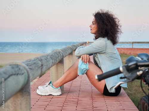 Girl looking at the horizon with a protective mask in hand, resting in a park, after a bike ride. Beach in the background.
