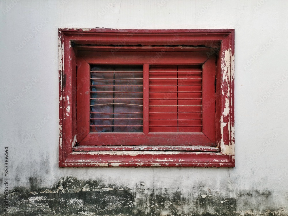 Old red wooden windows on a white cement wall with lemongrass stains. Antique wrought iron window design. Old red wooden window frames begin to decay.