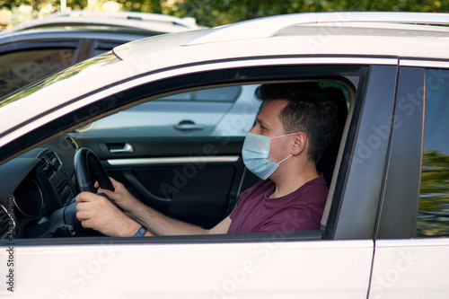 A man driving a car in a medical face mask during coronavirus outbreak, a taxi driver in a mask, protection from the virus. Quarantine, covid-19. © OneWellStudio