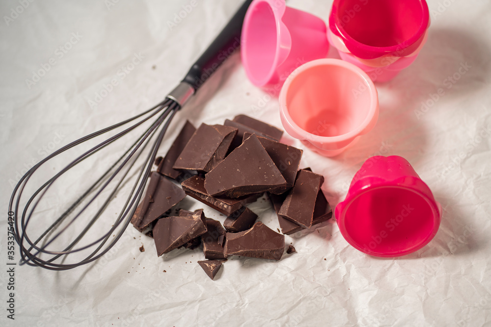 Fototapeta premium Getting ready for baking chocolate cupcakes, pink silicone forms and whisk with broken dark chocolate pieces on a baking paper
