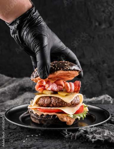 Chefs hand in black glove cooking the black burger with beaf steak, lettuce, tomatoes, slice of cheese, ham, pastrami and sauce on slate black background, close up photo