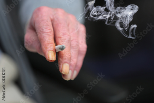The hand of an elderly person, holding a lit up cigarette. The nails of his index finger and ring finger are completely turned yellow because of the tar, nicotine and smoke. Concept of chain smoker.