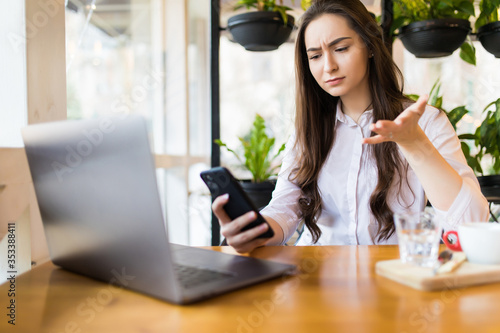 Worried woman talking on the phone trying to solve problems with multiple devices sitting in a desktop at cafe.