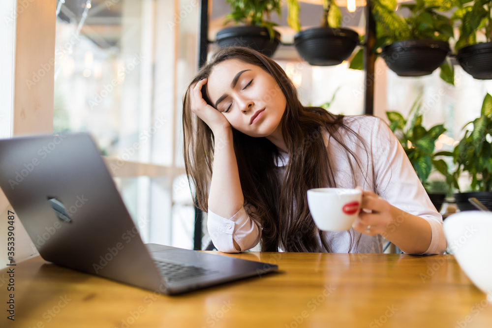 Portrait of a tired young woman sitting at the table with laptop computer while holding cup of coffee and sleeping at a cafe Portrait of a tired young woman sitting at the table with laptop computer while holding cup of coffee and sleeping at a cafe