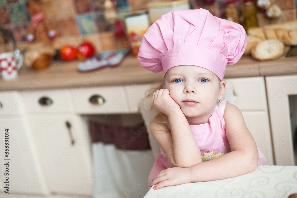 little girl in the kitchen in a pink chef's hat and apron sits at the table, pensive, dreamy. little girl in the kitchen in a pink chef's hat and apron sits at the table, pensive, dreamy.