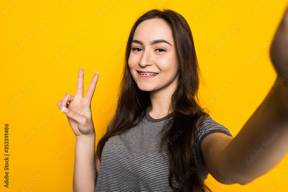 Portrait of stylish girl take selfie standing with peace sign in front of yellow wall. Portrait of stylish girl take selfie standing with peace sign in front of yellow wall.