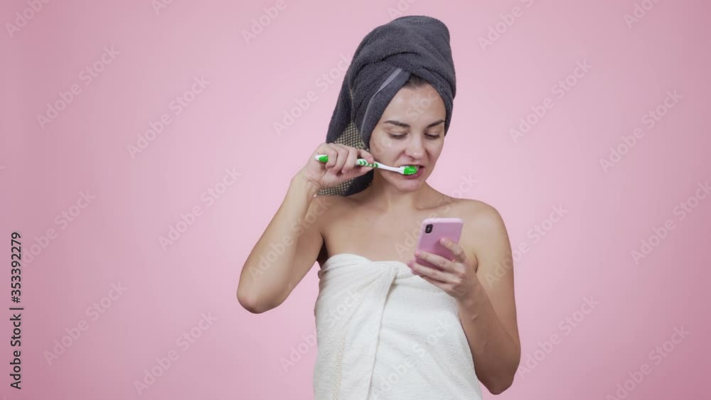 Girl in towel brushes her teeth and uses smartphone isolated on pink background Girl in towel brushes her teeth and uses smartphone isolated on pink background