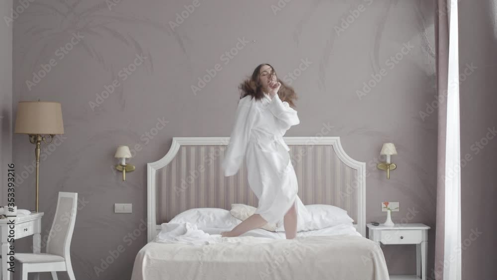 Funny young Caucasian woman jumping on bed in hotel room. Wide shot portrait of cheerful brunette girl in bathrobe having fun at luxurious resort in accommodations. Joy, leisure, lifestyle. Funny young Caucasian woman jumping on bed in hotel room. Wide shot portrait of cheerful brunette girl in bathrobe having fun at luxurious resort in accommodations. Joy, leisure, lifestyle.