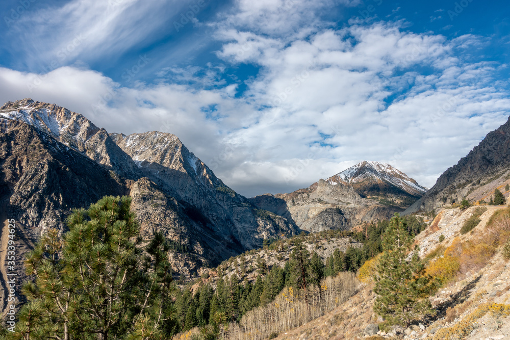 Fototapeta premium Dramatic clouds over snowy, high mountain peaks viewed from a valley filled with pine and aspen trees, Lundy Canyon, California