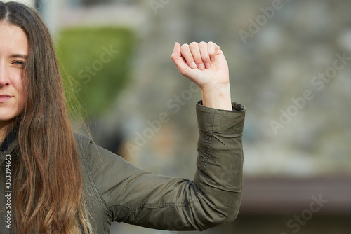 A half-part portrait of a young woman with long hair in a green jacket. A girl in an old street shows a fist as a symbol of the fight.