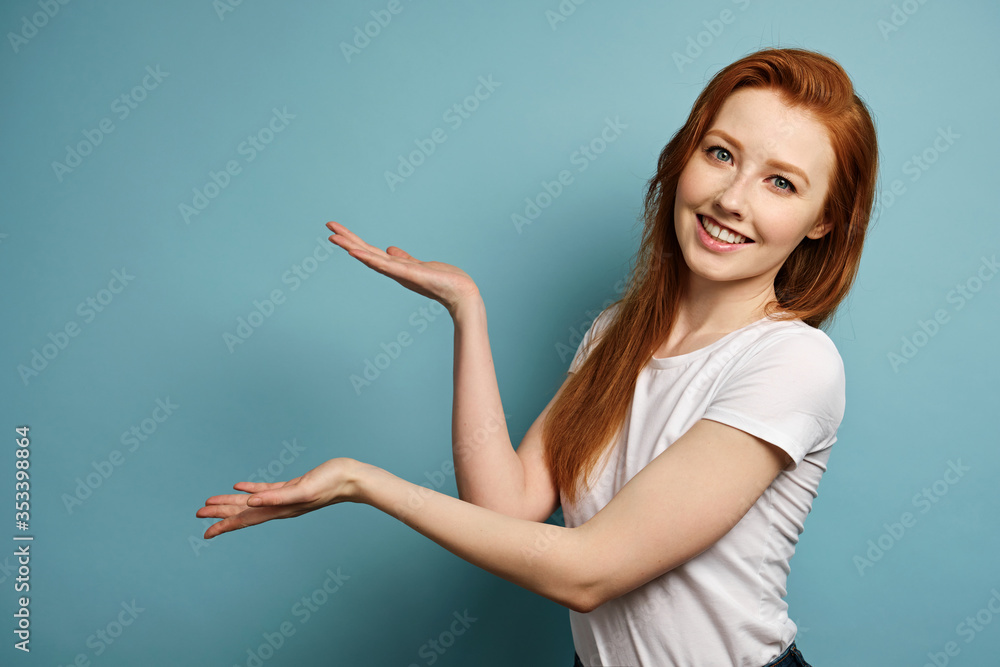 The red-haired girl stands on a blue background in a white T-shirt, smiles broadly and shows her palms to free space The red-haired girl stands on a blue background in a white T-shirt, smiles broadly and shows her palms to free space