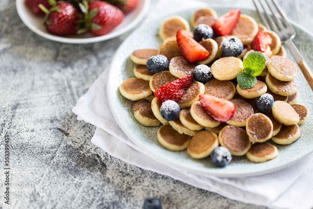 Tiny pancakes for breakfast. Cereal pancakes with blueberries, strawberries in plate on grey background. Trendy food. Selective focus Tiny pancakes for breakfast. Cereal pancakes with blueberries, strawberries in plate on grey background. Trendy food. Selective focus