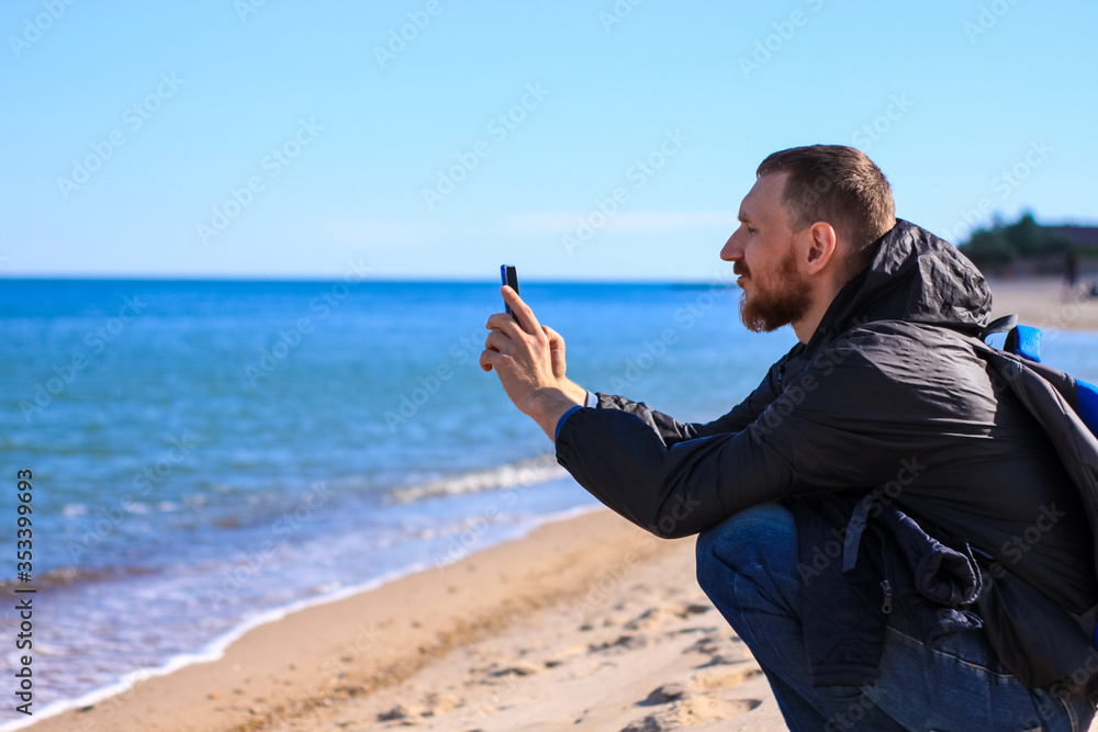 Bearded man in black raincoat on a blue sea landscape background with smartphone in hands talking. Middle aged guy calling friends on the cell phone on nature, people communication lifestyle concept. Bearded man in black raincoat on a blue sea landscape background with smartphone in hands talking. Middle aged guy calling friends on the cell phone on nature, people communication lifestyle concept.