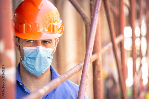 Ingegnere con casco arancio e camicia blu indossa una mascherina protettiva dentro un cantiere con delle impalcature in ferro photo