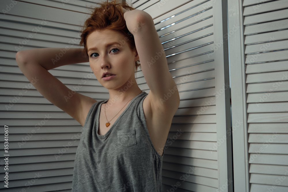 Redhead girl with freckles in a gray T-shirt against a white wooden screen, looking at the camera, collecting hair with hands up