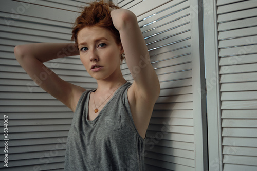 Redhead girl with freckles in a gray T-shirt against a white wooden screen, looking at the camera, collecting hair with hands up © bestsenny