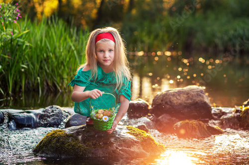 Fototapeta Naklejka Na Ścianę i Meble -  A girl in a green dress sits on a rock in the river. A blond kid with a red headband and red shoes is holding a basket. the river in the sun.