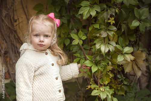 Little cute girl blonde in a light jacket and and a pink bow stands near the leaves of the vine and looks at the camera.