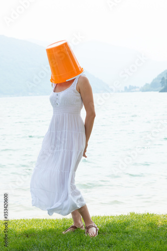Quadro su tela young woman standing by the lake with an orange bucket on her head