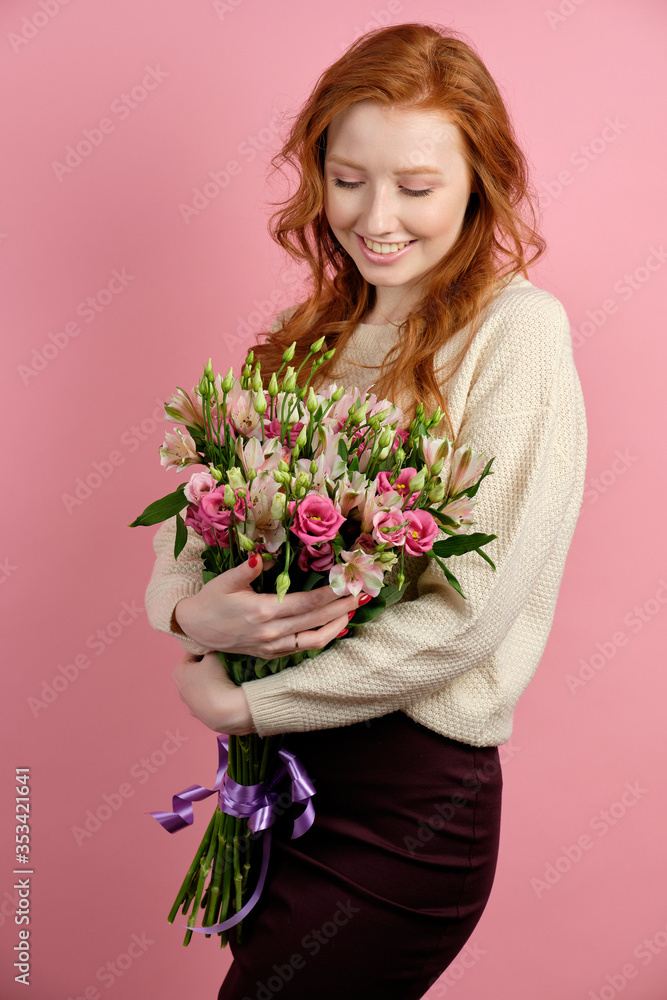 Beautiful red-haired girl stands on a pink background and looks at the bouquet in her hands with a smile.