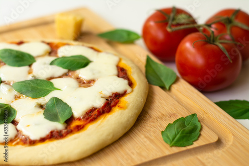 Homemade classic Italian Napoli pizza with tomato sauce, mozzarella cheese and basil leaves: pizza Napoletana. It is on a wooden cutting board surrounded by tomatoes, cheese and basil leaves