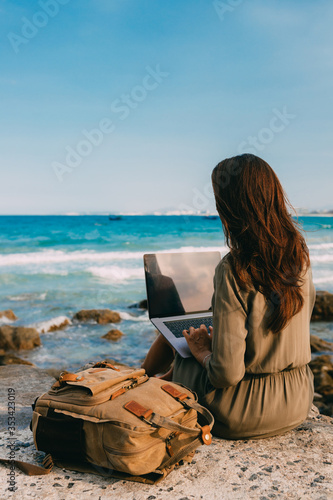 female freelancer sits with her back, works behind a laptop while traveling by the sea, remote work concept. Using modern technology, the Internet