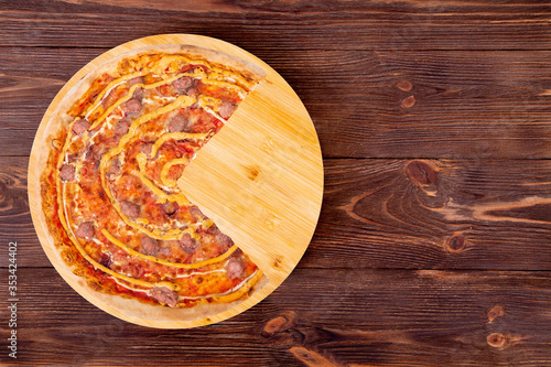Tasty pizza with beef sausages, mozzarella, various sauces and marinated red onions, without a quarter on a round wood platter which is on wooden rustic table, top view and space for text